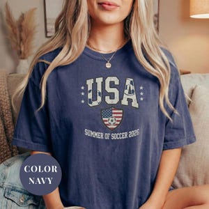 Navy Comfort Colors 1717 t-shirt with a vintage "USA Summer of Soccer 2026" graphic. Features bold varsity "USA" lettering with soccer ball patterns and a distressed American flag soccer shield. Patriotic apparel for World Cup 2026 fans.