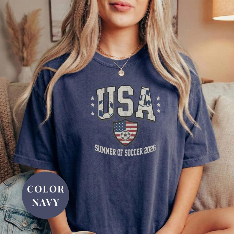 Navy Comfort Colors 1717 t-shirt with a vintage "USA Summer of Soccer 2026" graphic. Features bold varsity "USA" lettering with soccer ball patterns and a distressed American flag soccer shield. Patriotic apparel for World Cup 2026 fans.