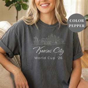 Pepper Comfort Colors T-shirt featuring a minimalist white line art Kansas City skyline design, elegant Kansas City script lettering, and World Cup ’26 text, styled as a soccer travel souvenir shirt for Kansas City fans, football lovers, and 2026 World Cup city apparel.