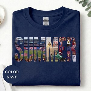 Navy Gildan t-shirt featuring a "SUMMER" photo collage graphic. The bold block letters are filled with USA soccer scenes, including a packed stadium, American flags, cheering fans, and a diving goalie. Patriotic sports apparel for World Cup 2026 supporters.