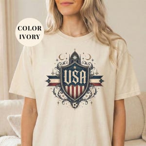 A woman wearing a white t-shirt with a graphic design featuring a shield with the text "USA" and various decorative elements.