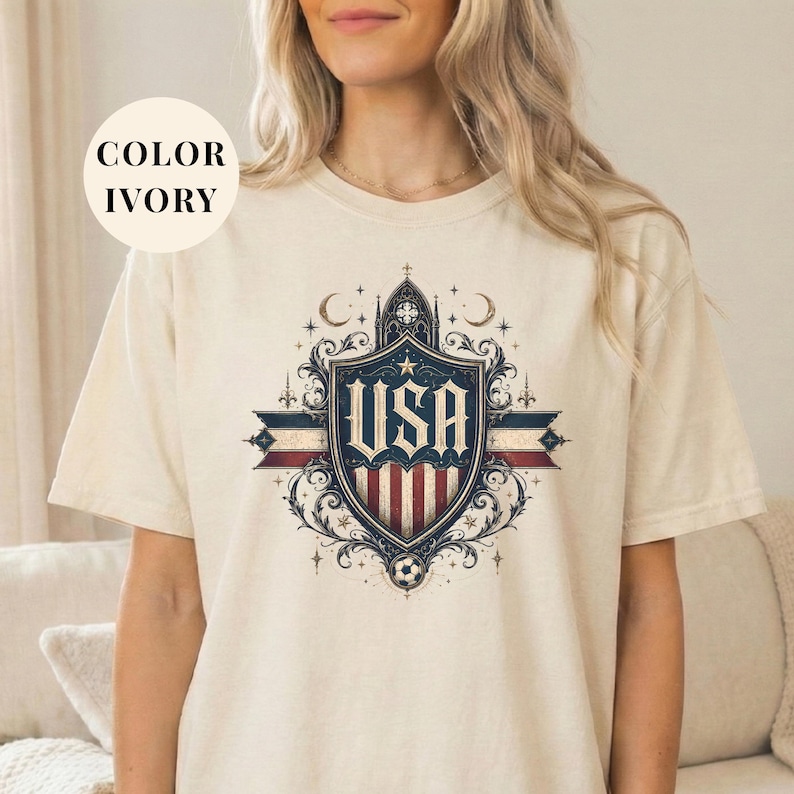 A woman wearing a white t-shirt with a graphic design featuring a shield with the text "USA" and various decorative elements.