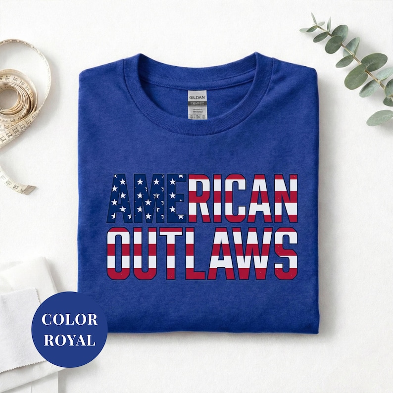 Royal t-shirt featuring a distressed retro design for "AMERICAN OUTLAWS". The bold collegiate block lettering is filled with a patriotic US national flag pattern of stars and stripes. High-quality vintage-style soccer apparel for passionate United States football supporters and tournament fans.