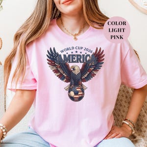 Pink t-shirt featuring a patriotic World Cup 2026 America design. A majestic bald eagle with red, white, and blue wings grasps a USA flag pattern soccer ball below "WORLD CUP 2026 AMERICA" text. Premium sports apparel for United States soccer fans and tournament supporters.