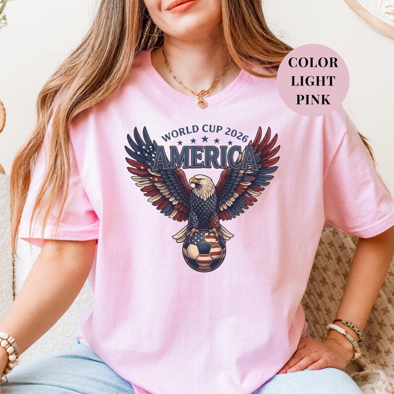Pink t-shirt featuring a patriotic World Cup 2026 America design. A majestic bald eagle with red, white, and blue wings grasps a USA flag pattern soccer ball below "WORLD CUP 2026 AMERICA" text. Premium sports apparel for United States soccer fans and tournament supporters.