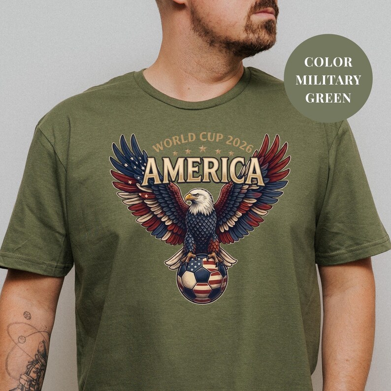 Green t-shirt featuring a patriotic World Cup 2026 America design. A majestic bald eagle with red, white, and blue wings grasps a USA flag pattern soccer ball below "WORLD CUP 2026 AMERICA" text. Premium sports apparel for United States soccer fans and tournament supporters.