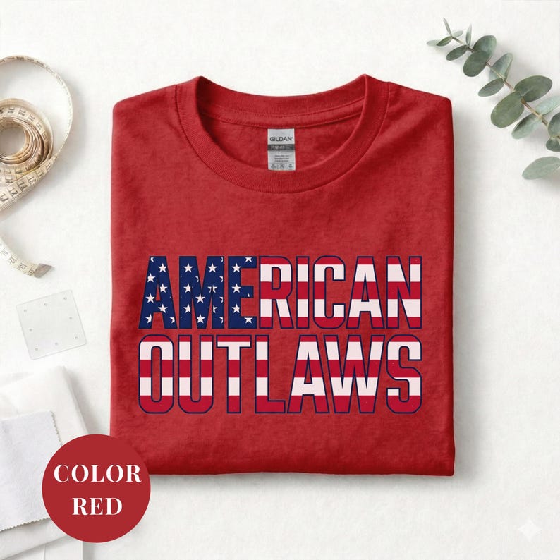 Red t-shirt featuring a distressed retro design for "AMERICAN OUTLAWS". The bold collegiate block lettering is filled with a patriotic US national flag pattern of stars and stripes. High-quality vintage-style soccer apparel for passionate United States football supporters and tournament fans.