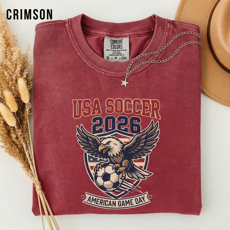 May include: Crimson red t-shirt with a graphic featuring an eagle clutching a soccer ball, the text "USA SOCCER 2026", and "AMERICAN GAME DAY". A silver necklace with a star pendant and a brown hat are also visible.