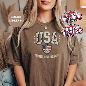 Espresso brown Comfort Colors 1717 t-shirt with a vintage "USA Summer of Soccer 2026" graphic. Features bold varsity "USA" lettering with soccer ball patterns and a distressed American flag soccer shield. Patriotic apparel for World Cup 2026 fans.