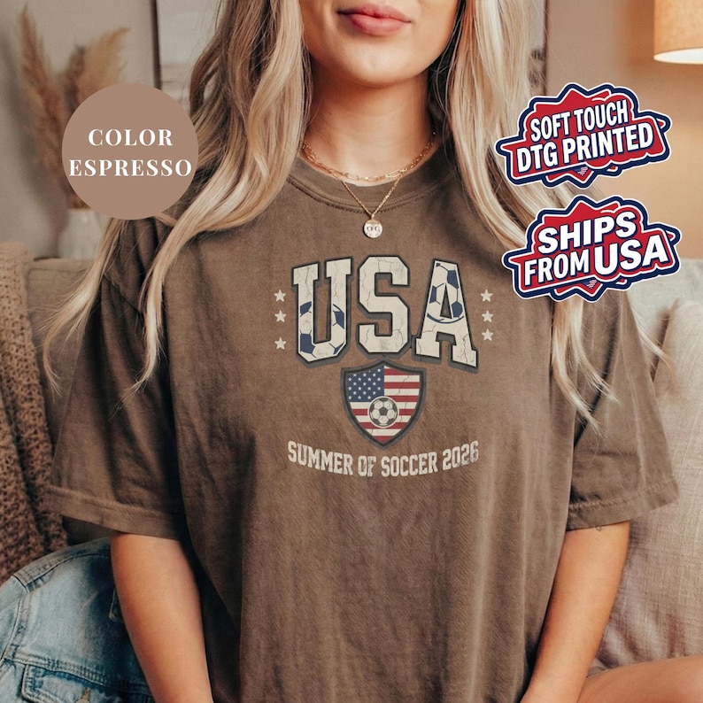 Espresso brown Comfort Colors 1717 t-shirt with a vintage "USA Summer of Soccer 2026" graphic. Features bold varsity "USA" lettering with soccer ball patterns and a distressed American flag soccer shield. Patriotic apparel for World Cup 2026 fans.