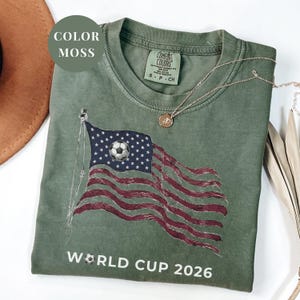 Moss Comfort Colors 1717 t-shirt featuring a vintage-style waving American flag. The design includes a soccer ball integrated into the stars and "WORLD CUP 2026" bold text. High-quality patriotic sports apparel for United States soccer fans and tournament supporters.
