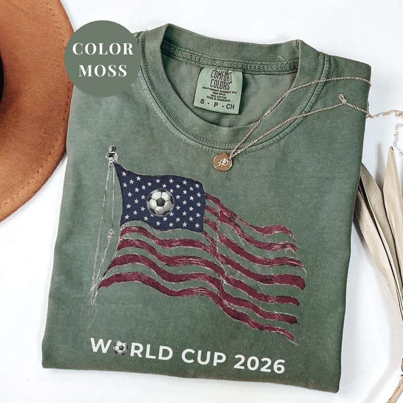 Moss Comfort Colors 1717 t-shirt featuring a vintage-style waving American flag. The design includes a soccer ball integrated into the stars and "WORLD CUP 2026" bold text. High-quality patriotic sports apparel for United States soccer fans and tournament supporters.