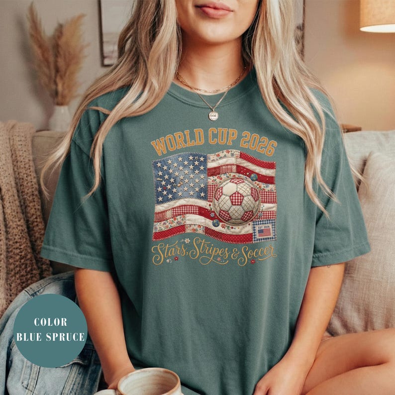 Blue spruce colored t-shirt featuring a vintage patchwork American flag design for World Cup 2026. The faux quilted graphic includes floral and gingham fabric textures, a stitched soccer ball, and "Stars, Stripes & Soccer" text. Unique patriotic sports apparel for women and USA soccer fans.