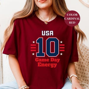 A woman wearing a maroon t-shirt with the text "USA 10 Game Day Energy" printed on it.