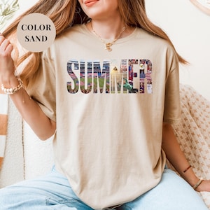 Sand Gildan t-shirt featuring a "SUMMER" photo collage graphic. The bold block letters are filled with USA soccer scenes, including a packed stadium, American flags, cheering fans, and a diving goalie. Patriotic sports apparel for World Cup 2026 supporters.