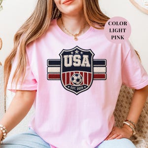 A woman wearing a pink t-shirt with a shield-shaped design featuring the text "USA" and a soccer ball, along with the year "EST. 2023".