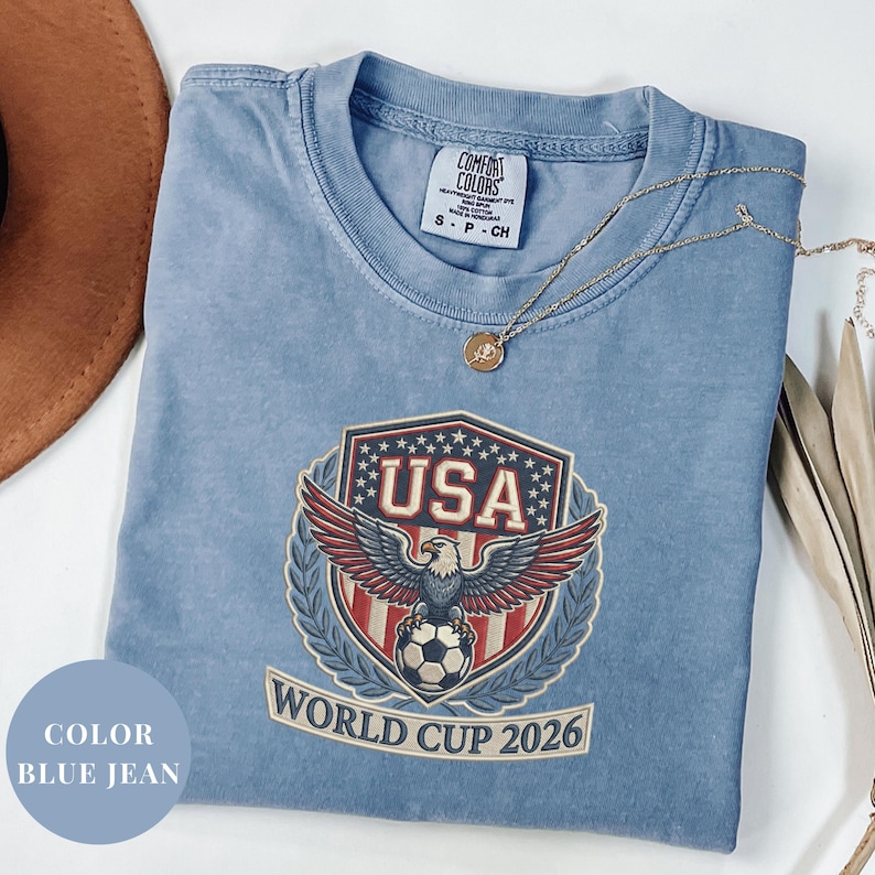 Blue Comfort Colors 1717 t-shirt featuring a detailed faux embroidery USA World Cup 2026 emblem. The crest shows a bald eagle perched on a soccer ball, surrounded by a laurel wreath and stars. Premium patriotic soccer fan apparel for World Cup 2026.