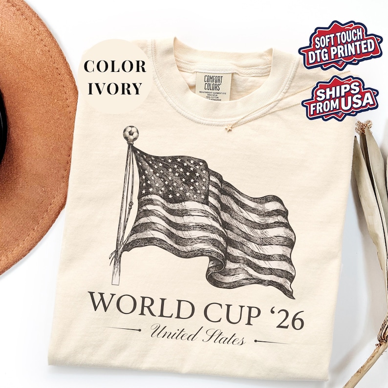 Ivory Comfort Colors 1717 t-shirt featuring a vintage hand-drawn sketch of a waving American flag. Includes "WORLD CUP '26" and "United States" text, with a subtle soccer ball detail on the flagpole. High-quality patriotic soccer apparel for the 2026 world tournament fans.