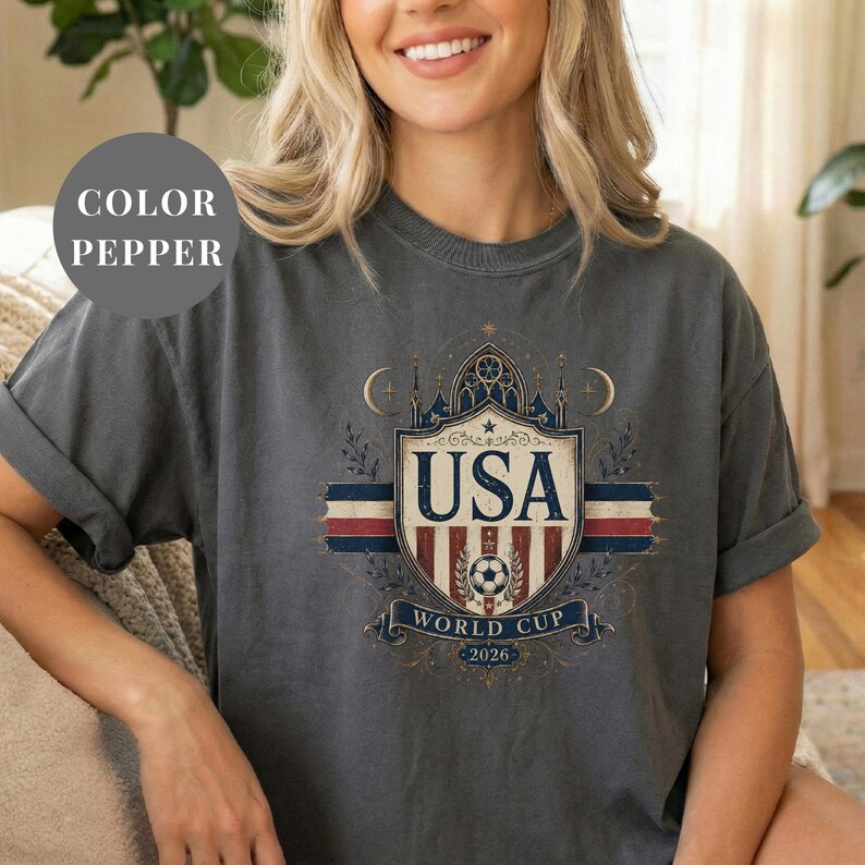 A woman wearing a gray t-shirt with a graphic design featuring the text "USA WORLD CUP 2026" and a shield-shaped emblem.
