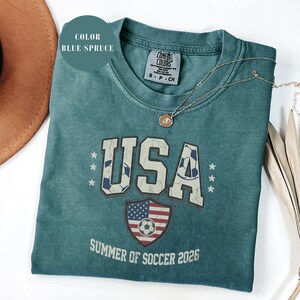 Blue spruce Comfort Colors 1717 t-shirt with a vintage "USA Summer of Soccer 2026" graphic. Features bold varsity "USA" lettering with soccer ball patterns and a distressed American flag soccer shield. Patriotic apparel for World Cup 2026 fans.