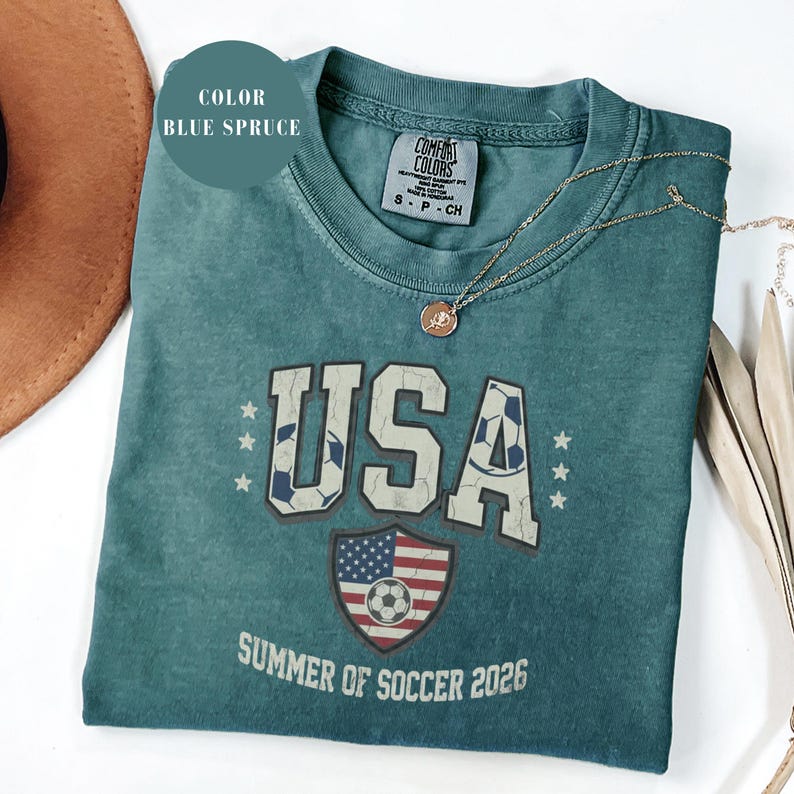 Blue spruce Comfort Colors 1717 t-shirt with a vintage "USA Summer of Soccer 2026" graphic. Features bold varsity "USA" lettering with soccer ball patterns and a distressed American flag soccer shield. Patriotic apparel for World Cup 2026 fans.