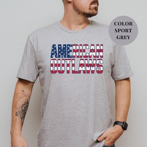 Sport Grey t-shirt featuring a distressed retro design for "AMERICAN OUTLAWS". The bold collegiate block lettering is filled with a patriotic US national flag pattern of stars and stripes. High-quality vintage-style soccer apparel for passionate United States football supporters and tournament fans.
