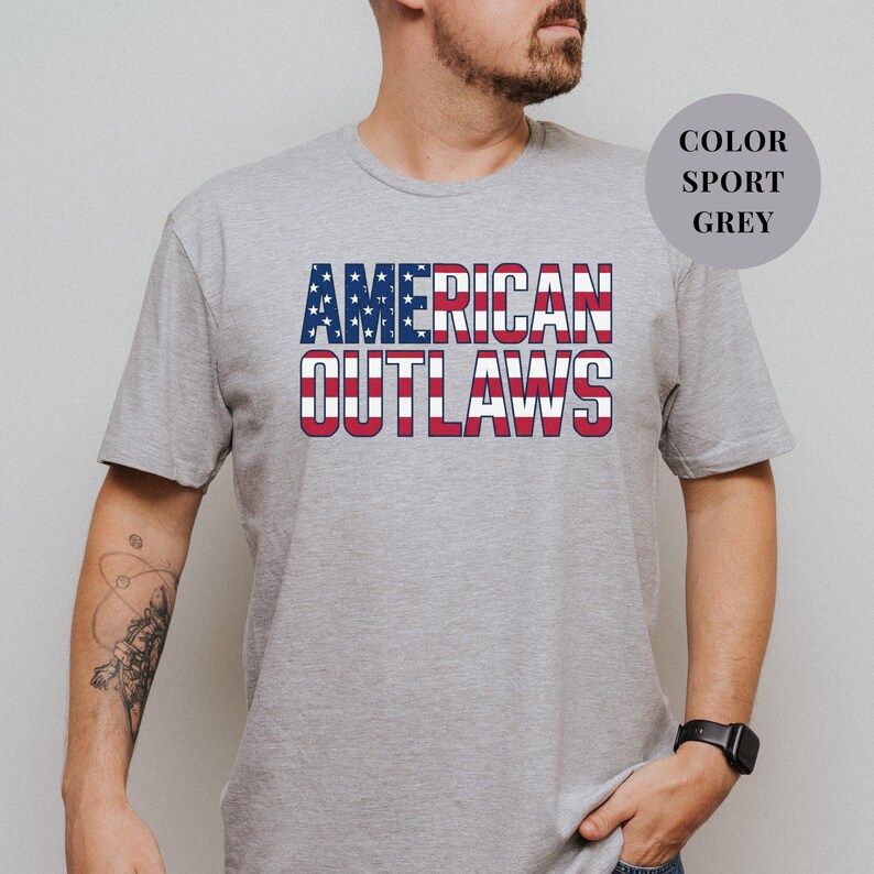 Sport Grey t-shirt featuring a distressed retro design for "AMERICAN OUTLAWS". The bold collegiate block lettering is filled with a patriotic US national flag pattern of stars and stripes. High-quality vintage-style soccer apparel for passionate United States football supporters and tournament fans.