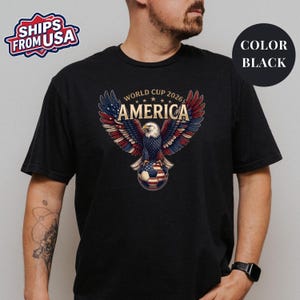 May include: A black short-sleeved t-shirt featuring a graphic of a bald eagle with wings spread wide, perched on a soccer ball. The eagle's wings are colored in red, white, and blue. The text reads WORLD CUP 2026 AMERICA above the eagle.