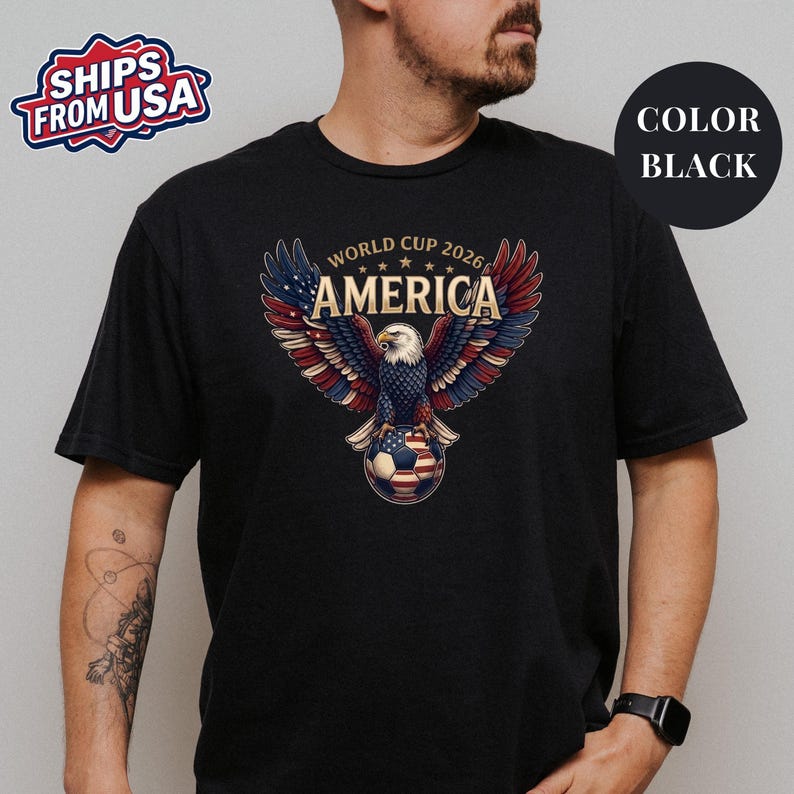 May include: A black short-sleeved t-shirt featuring a graphic of a bald eagle with wings spread wide, perched on a soccer ball. The eagle's wings are colored in red, white, and blue. The text reads WORLD CUP 2026 AMERICA above the eagle.