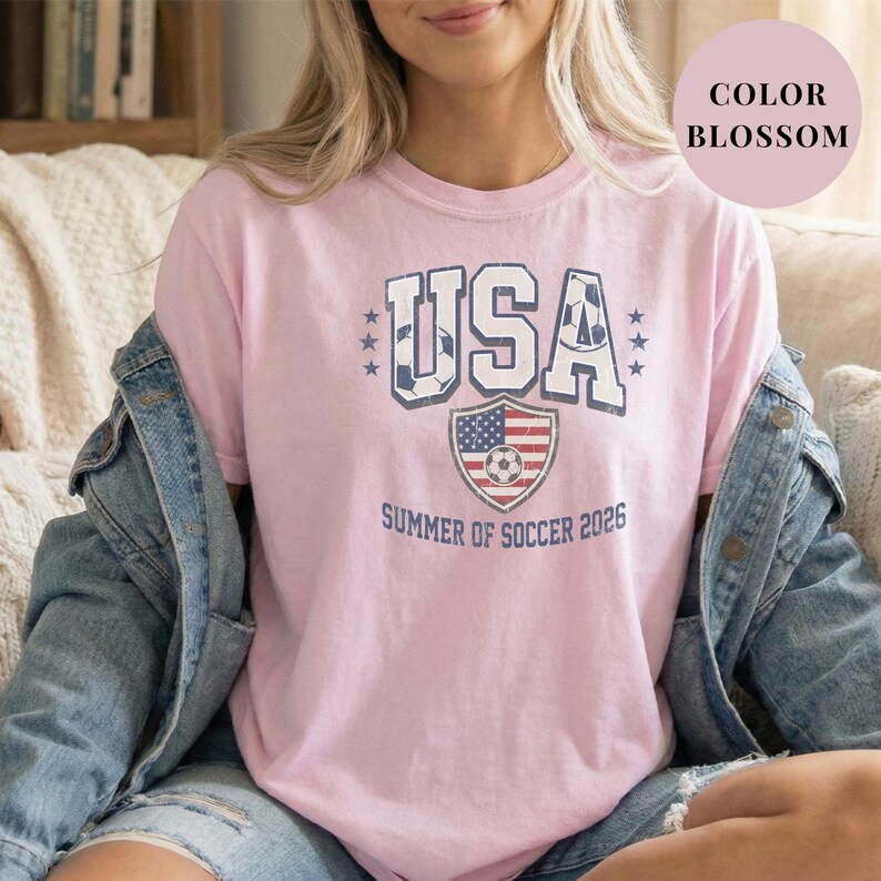 Blossom pink Comfort Colors 1717 t-shirt with a vintage "USA Summer of Soccer 2026" graphic. Features bold varsity "USA" lettering with soccer ball patterns and a distressed American flag soccer shield. Patriotic apparel for World Cup 2026 fans.