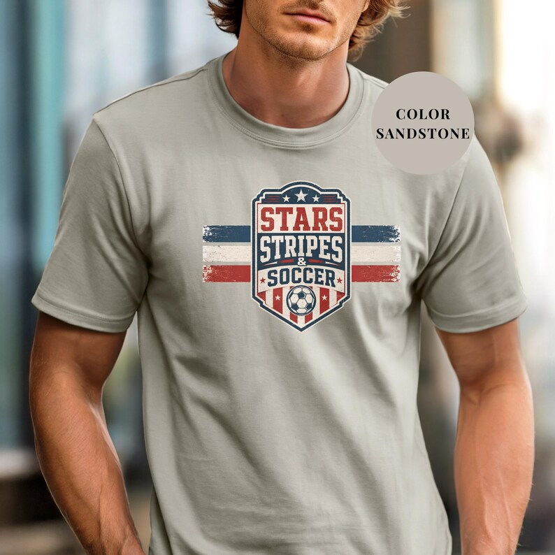 A man wearing a gray t-shirt with a graphic design featuring a shield-shaped logo with the text "STARS STRIPS SOCCER" and an American flag pattern.