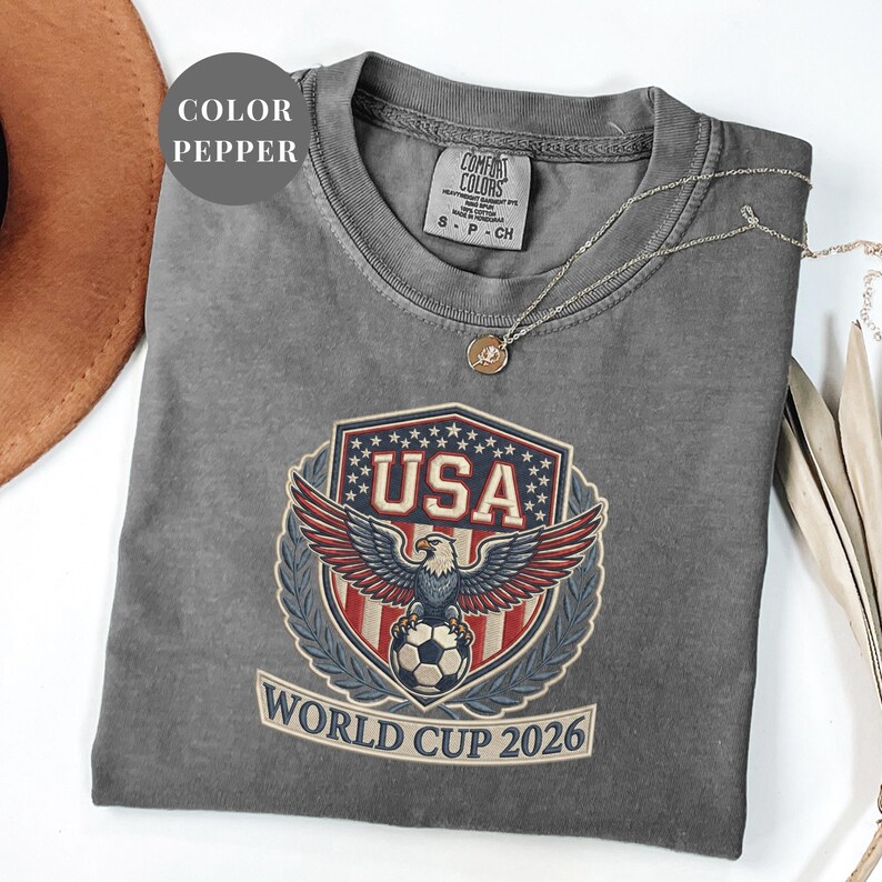 Pepper Comfort Colors 1717 t-shirt featuring a detailed faux embroidery USA World Cup 2026 emblem. The crest shows a bald eagle perched on a soccer ball, surrounded by a laurel wreath and stars. Premium patriotic soccer fan apparel for World Cup 2026.