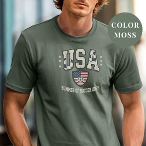 Moss Comfort Colors 1717 t-shirt with a vintage "USA Summer of Soccer 2026" graphic. Features bold varsity "USA" lettering with soccer ball patterns and a distressed American flag soccer shield. Patriotic apparel for World Cup 2026 fans.