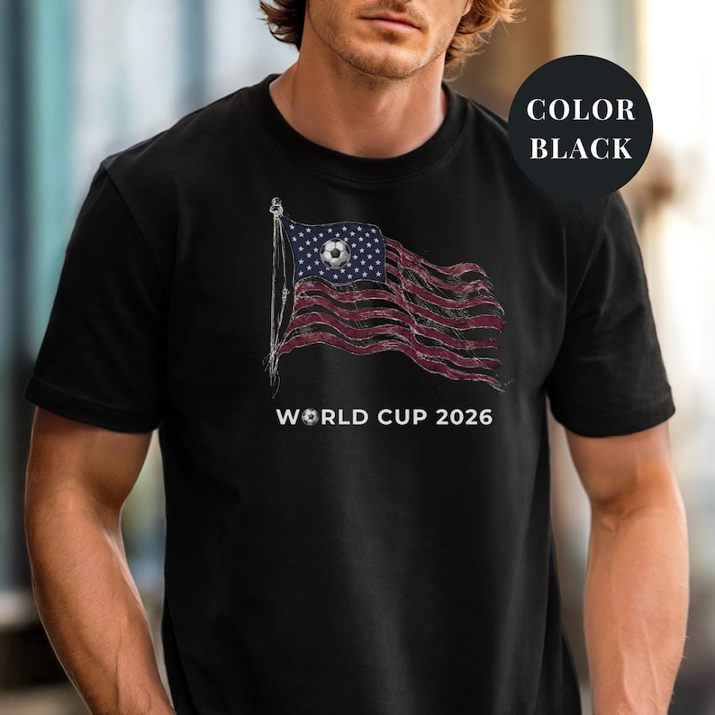 Black Comfort Colors 1717 t-shirt featuring a vintage-style waving American flag. The design includes a soccer ball integrated into the stars and "WORLD CUP 2026" bold text. High-quality patriotic sports apparel for United States soccer fans and tournament supporters.