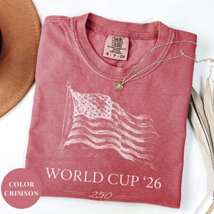 Crimson Comfort Colors 1717 t-shirt featuring a minimalist vintage sketch of a waving American flag. Includes "WORLD CUP '26" and a stylized "250" text celebrating the USA Semiquincentennial. Premium patriotic sports apparel for United States soccer fans and 2026 host nation supporters.
