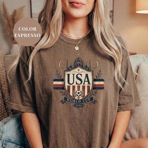 A woman wearing a brown t-shirt with a graphic design featuring the text "USA WORLD CUP 2026" and a shield-shaped emblem.