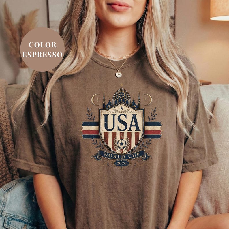 A woman wearing a brown t-shirt with a graphic design featuring the text "USA WORLD CUP 2026" and a shield-shaped emblem.