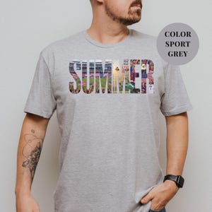 Grey Gildan t-shirt featuring a "SUMMER" photo collage graphic. The bold block letters are filled with USA soccer scenes, including a packed stadium, American flags, cheering fans, and a diving goalie. Patriotic sports apparel for World Cup 2026 supporters.