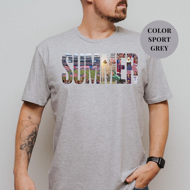 Grey Gildan t-shirt featuring a "SUMMER" photo collage graphic. The bold block letters are filled with USA soccer scenes, including a packed stadium, American flags, cheering fans, and a diving goalie. Patriotic sports apparel for World Cup 2026 supporters.
