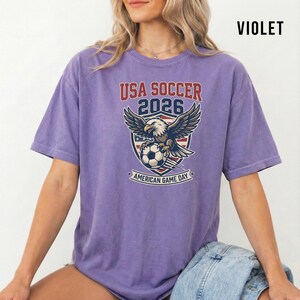May include: Violet-colored t-shirt featuring a graphic for the 2026 USA Soccer American Game Day. The design includes an eagle, soccer ball, and the text "USA SOCCER 2026".