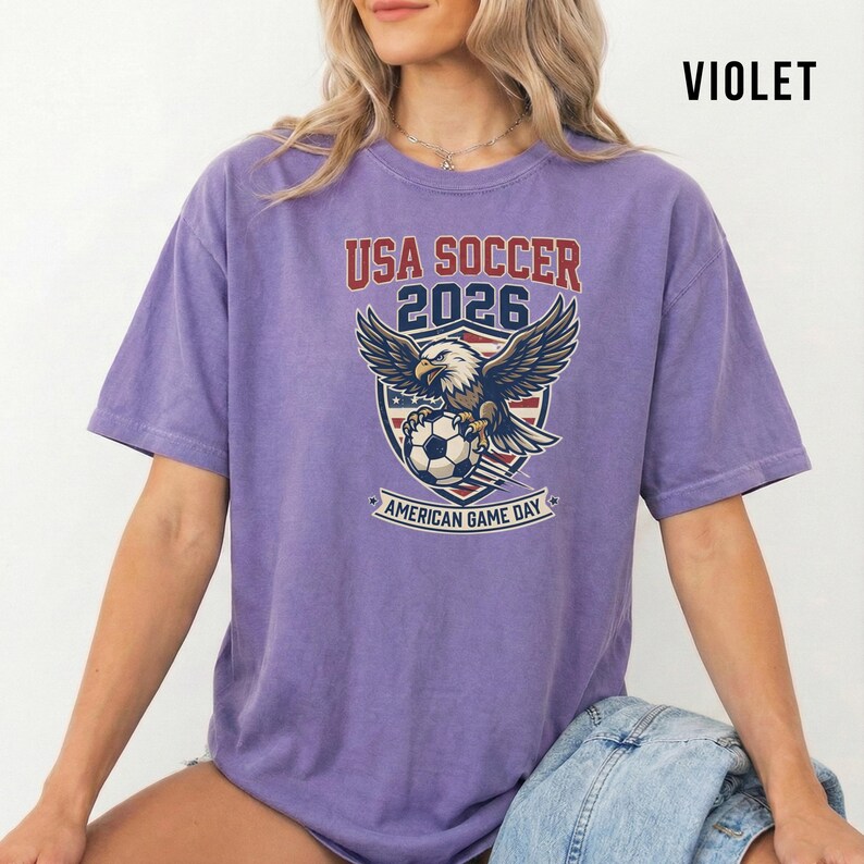 May include: Violet-colored t-shirt featuring a graphic for the 2026 USA Soccer American Game Day. The design includes an eagle, soccer ball, and the text "USA SOCCER 2026".