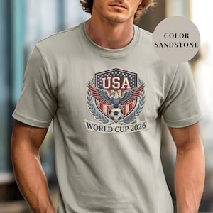 Sandstone Comfort Colors 1717 t-shirt featuring a detailed faux embroidery USA World Cup 2026 emblem. The crest shows a bald eagle perched on a soccer ball, surrounded by a laurel wreath and stars. Premium patriotic soccer fan apparel for World Cup 2026.