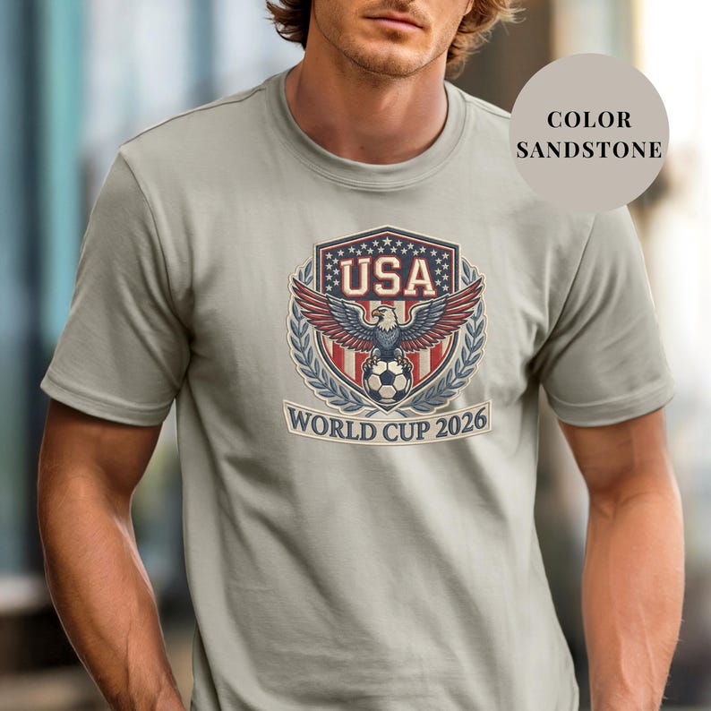 Sandstone Comfort Colors 1717 t-shirt featuring a detailed faux embroidery USA World Cup 2026 emblem. The crest shows a bald eagle perched on a soccer ball, surrounded by a laurel wreath and stars. Premium patriotic soccer fan apparel for World Cup 2026.
