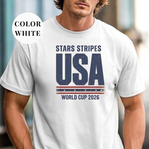 White t-shirt featuring a distressed vintage "STARS STRIPES USA WORLD CUP 2026" graphic. The design includes weathered block lettering for "USA" and a patriotic red, white, and blue banner with stars. High-quality sports apparel for United States soccer fans and world tournament supporters.