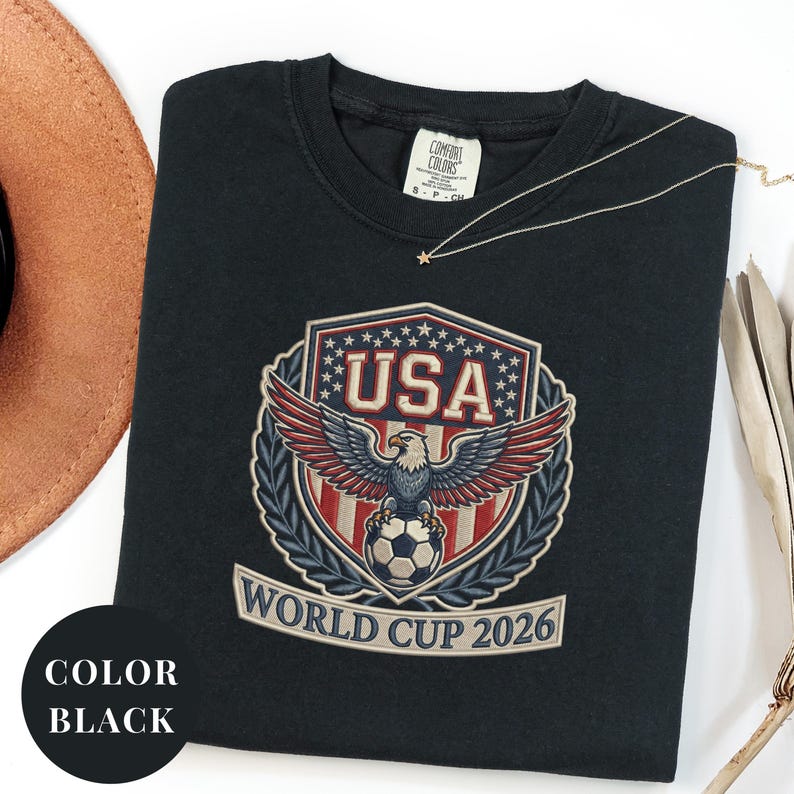 Black Comfort Colors 1717 t-shirt featuring a detailed faux embroidery USA World Cup 2026 emblem. The crest shows a bald eagle perched on a soccer ball, surrounded by a laurel wreath and stars. Premium patriotic soccer fan apparel for World Cup 2026.
