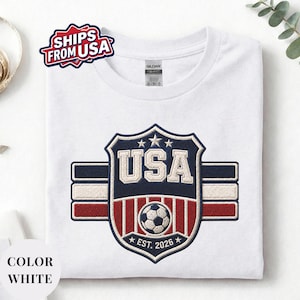 A white t-shirt with a large embroidered patch that says "USA" and features a soccer ball and stars, along with the text "EST. 2026".