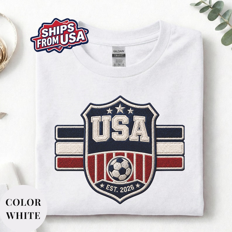 A white t-shirt with a large embroidered patch that says "USA" and features a soccer ball and stars, along with the text "EST. 2026".