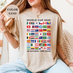 Sand t-shirt featuring a comprehensive grid of 48 national flags for the World Cup 2026. The design includes bold "WORLD CUP 2026" text at the top and the slogan "48 NATIONS • ONE GAME" at the bottom. Premium international soccer fan apparel for the upcoming world tournament in North America.