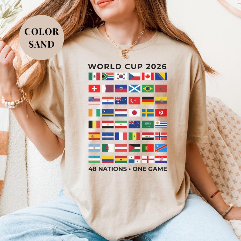 Sand t-shirt featuring a comprehensive grid of 48 national flags for the World Cup 2026. The design includes bold "WORLD CUP 2026" text at the top and the slogan "48 NATIONS • ONE GAME" at the bottom. Premium international soccer fan apparel for the upcoming world tournament in North America.