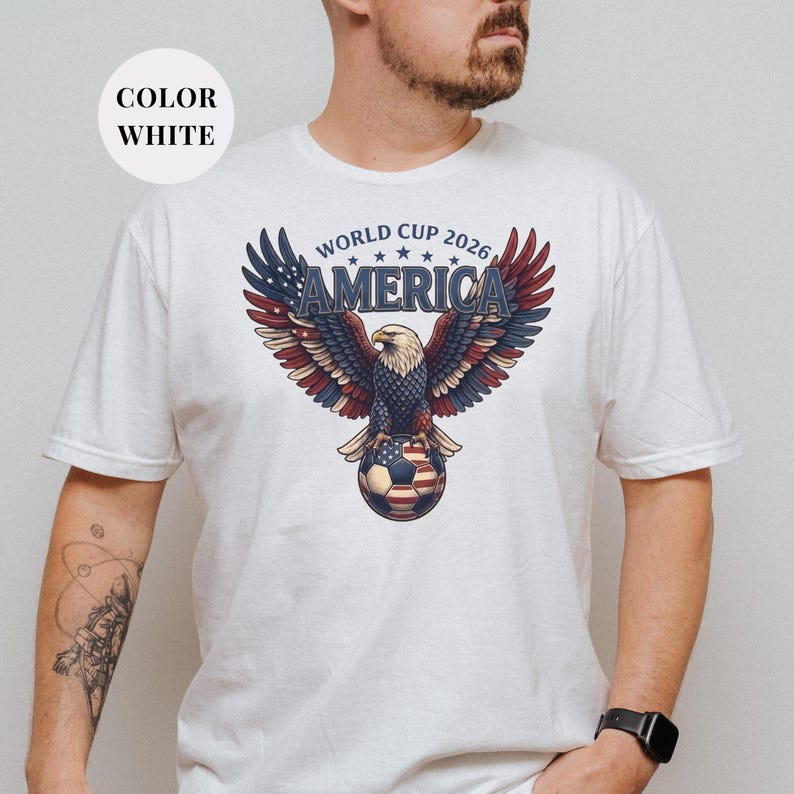 White t-shirt featuring a patriotic World Cup 2026 America design. A majestic bald eagle with red, white, and blue wings grasps a USA flag pattern soccer ball below "WORLD CUP 2026 AMERICA" text. Premium sports apparel for United States soccer fans and tournament supporters.
